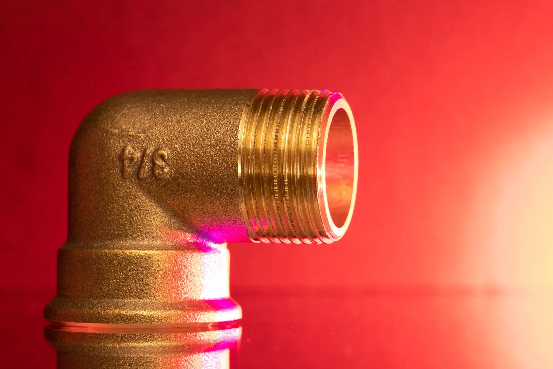 Brass elbow fitting lit hard against a deep red background, threads sharp