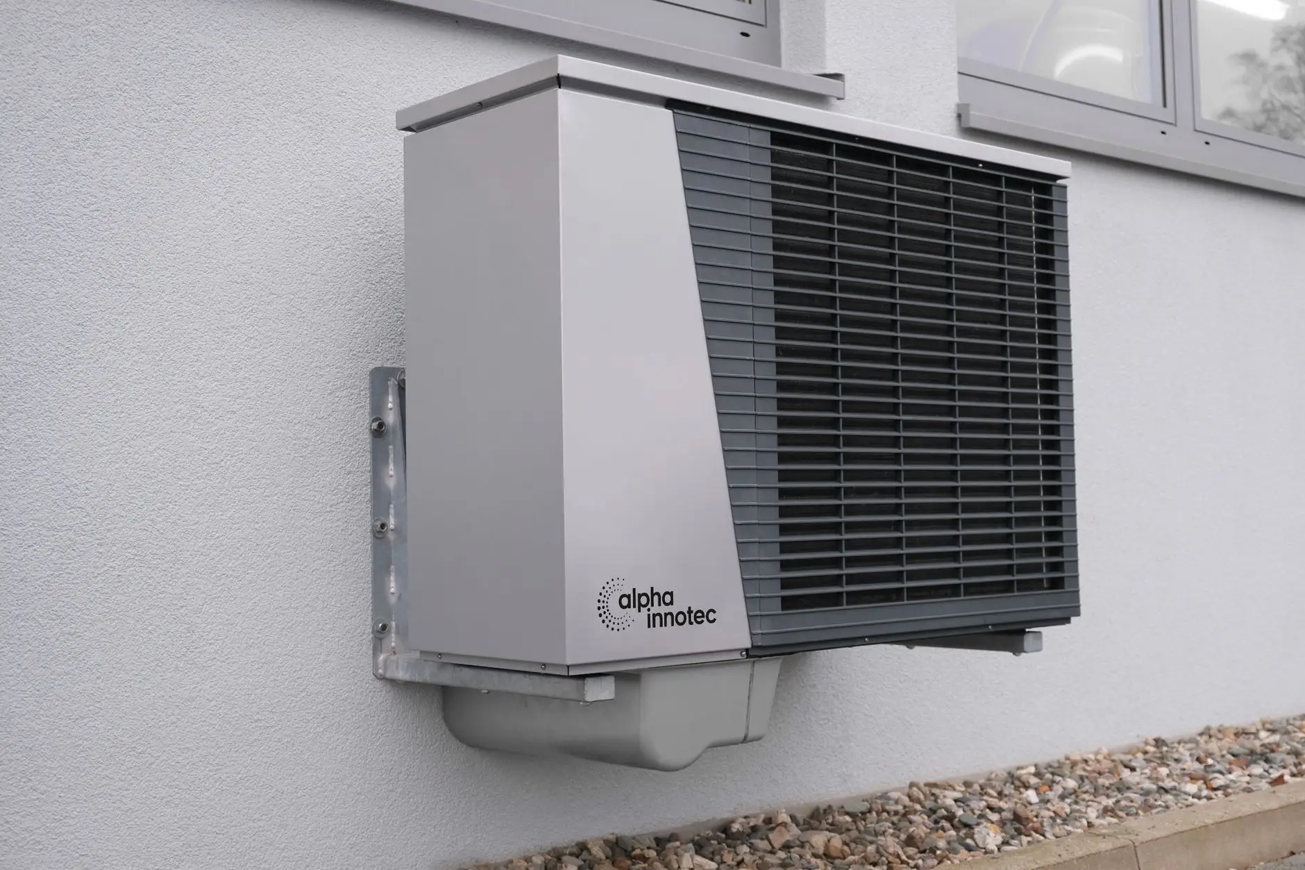 Heat pump unit mounted on a pale stucco wall above a gravel strip