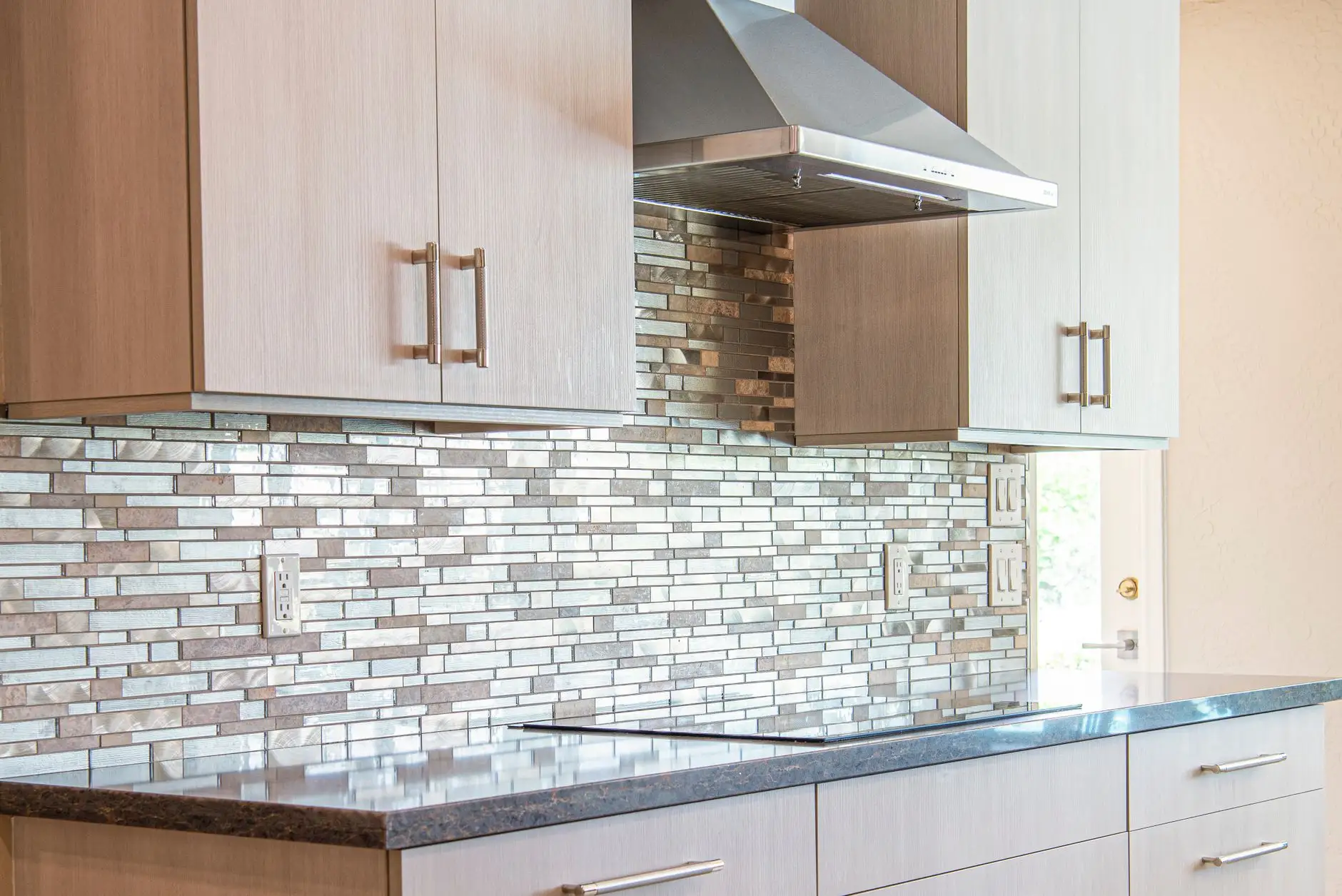 Glass mosaic backsplash and dark stone counter under a stainless range hood