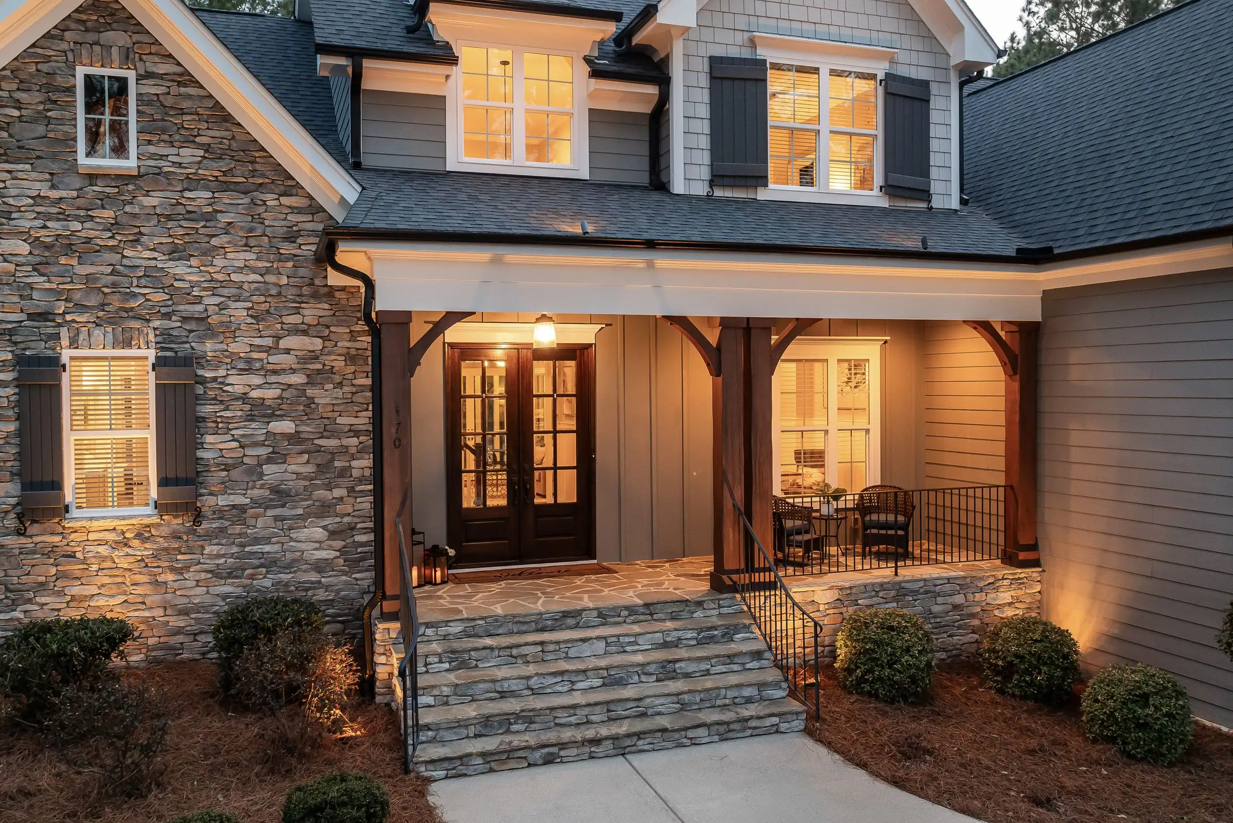 Stone and shingle house front lit from within at dusk, deep covered porch