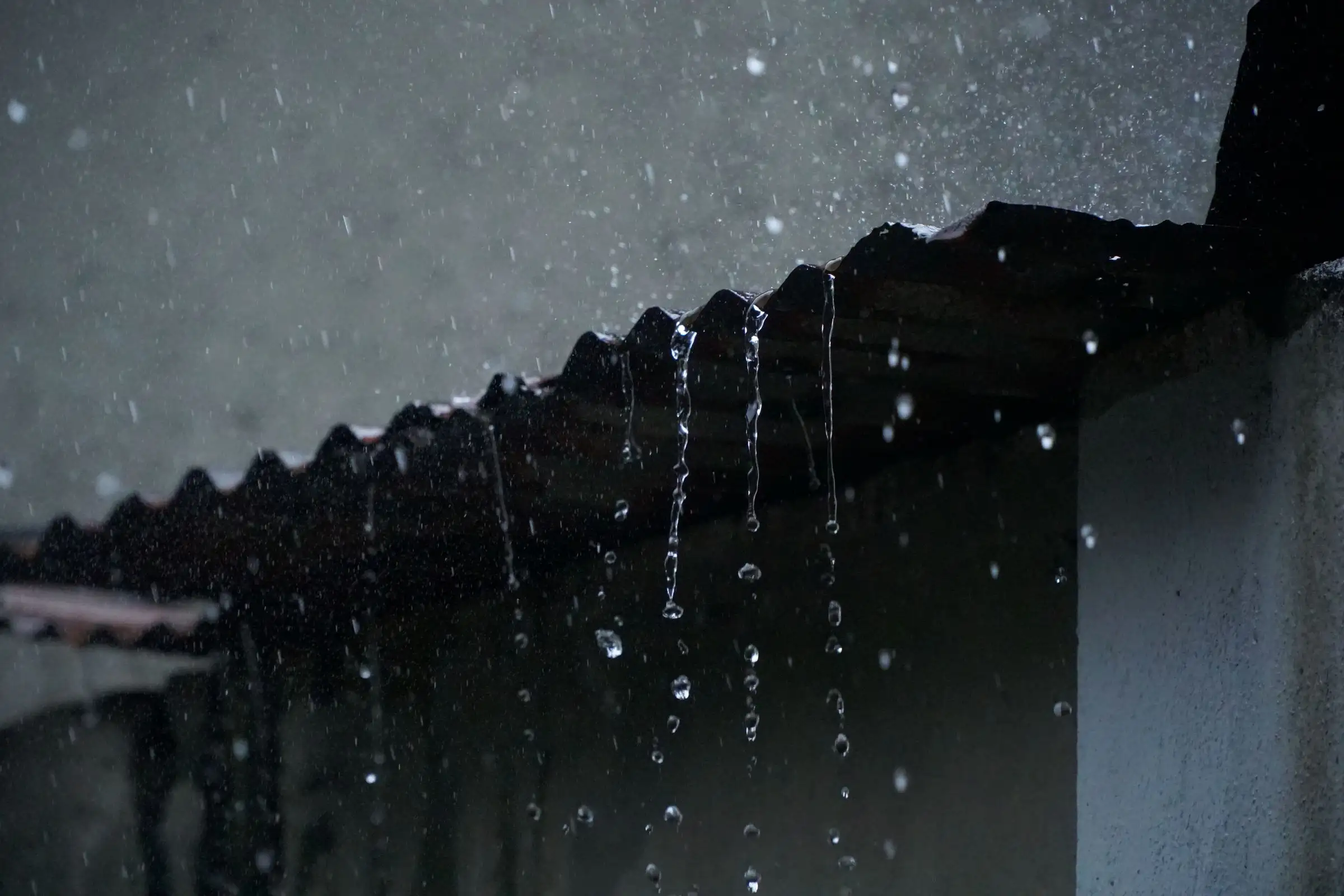 Rain running off the edge of a roof in low light, water breaking into strands as it falls