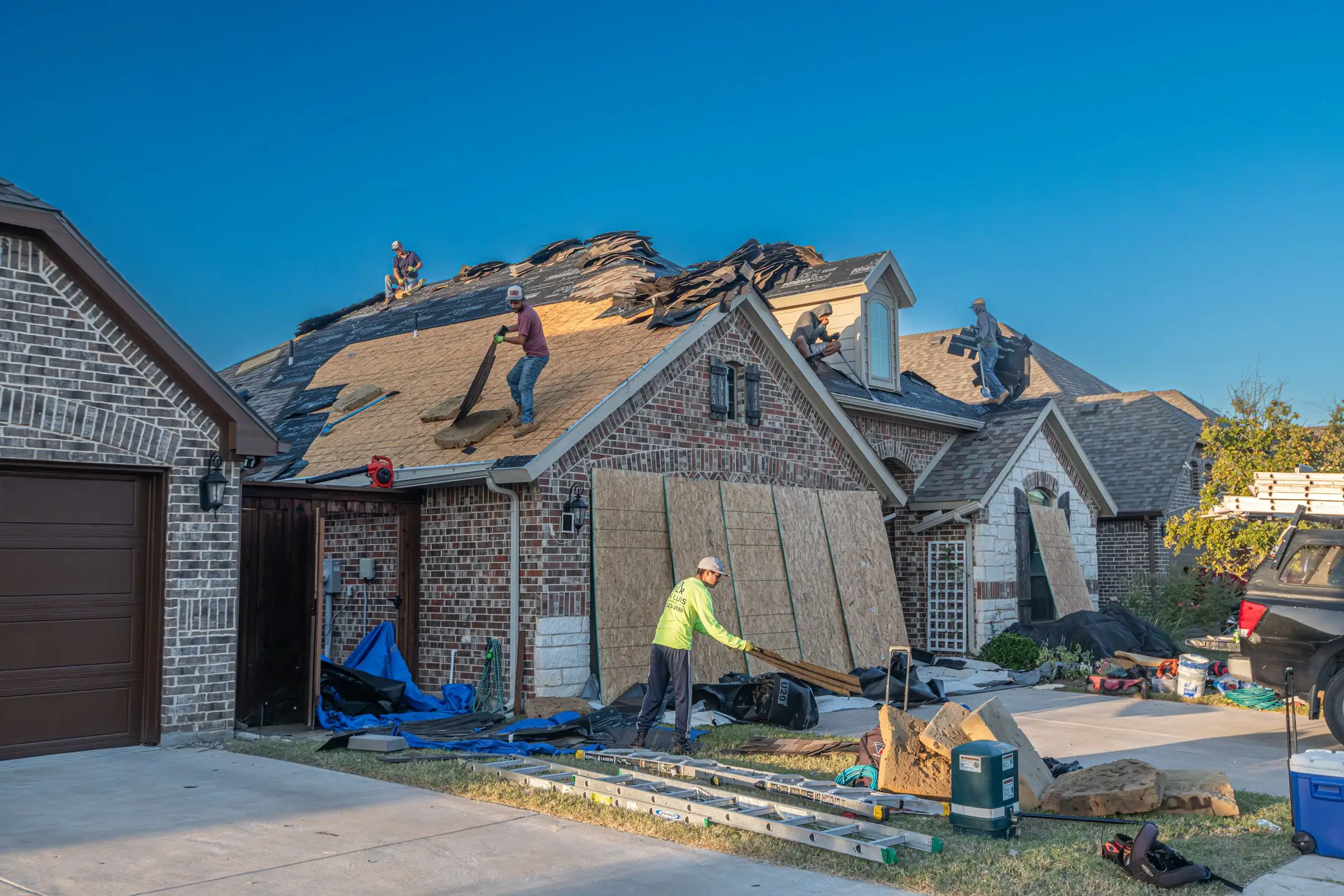Roofing crew stripping old shingles off a brick house, bare decking and debris across the slope