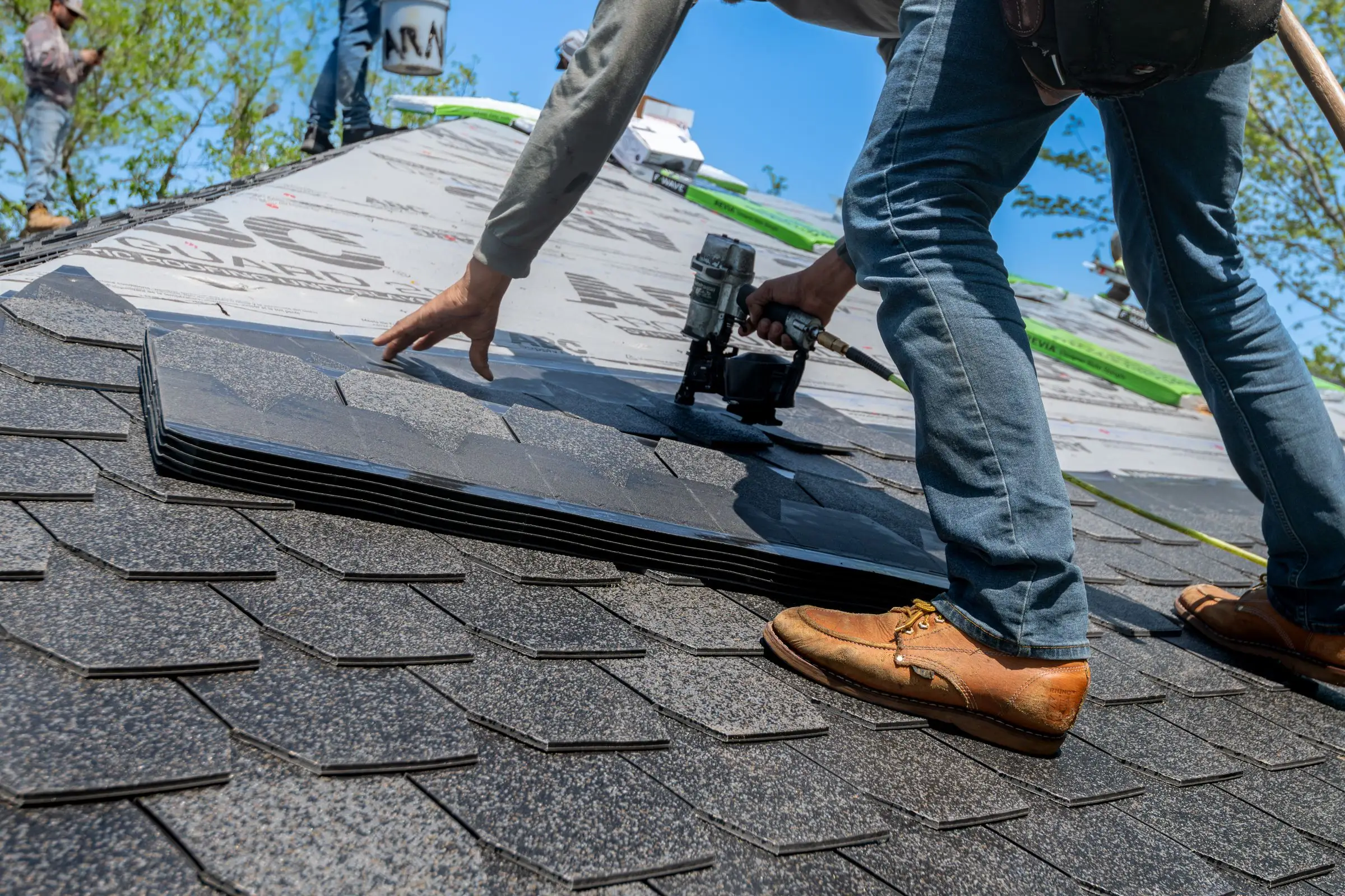 Roofer nailing a course of architectural asphalt shingles up a steep slope, bare underlayment above
